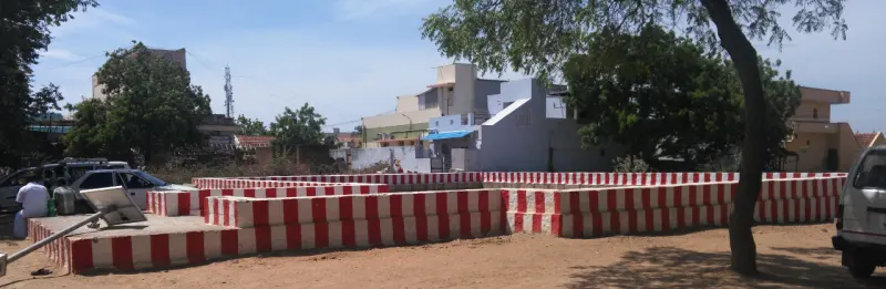 Low walls painted in red and white stripes enclose an open sandy area, with houses and trees beyond.