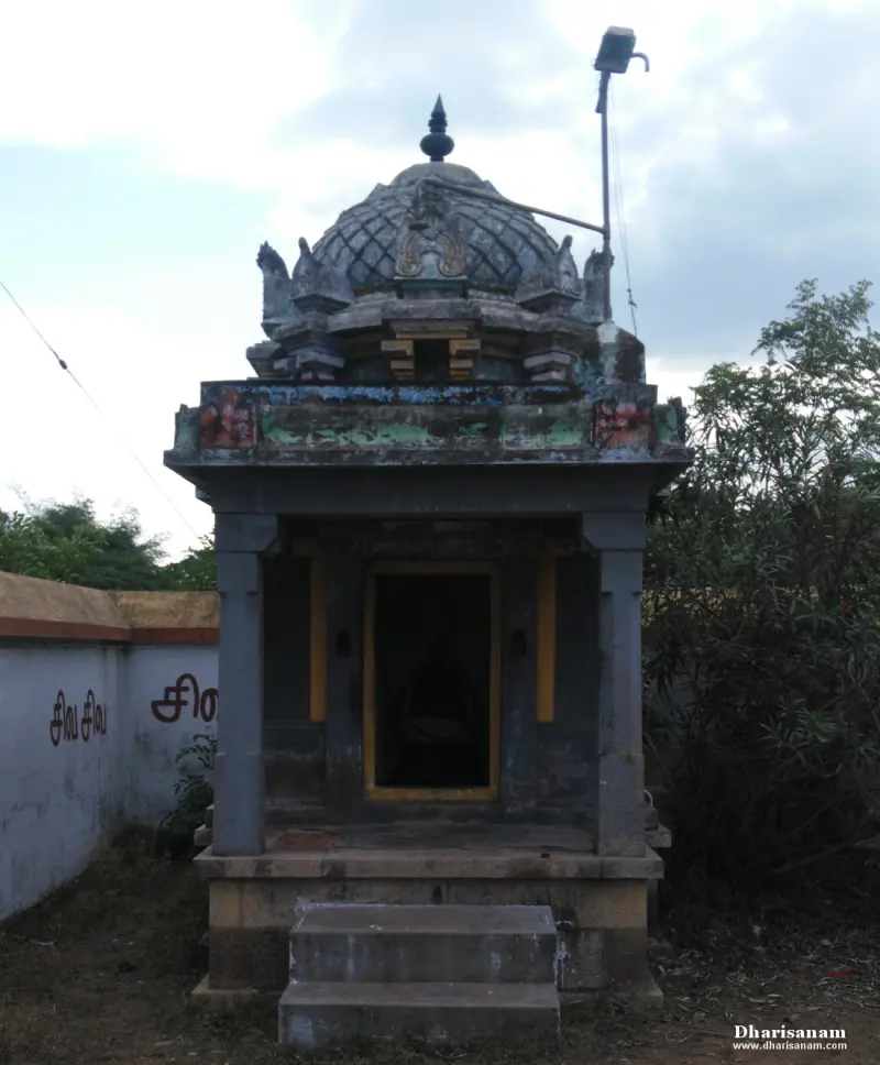 A small stone shrine with a domed roof, standing beside a wall with Tamil letters.