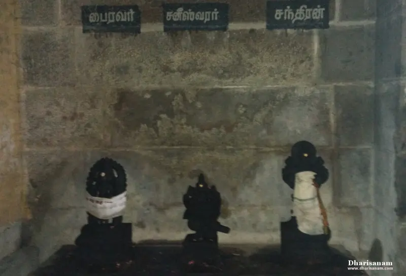Three dark idols under Tamil name boards against a stone wall
