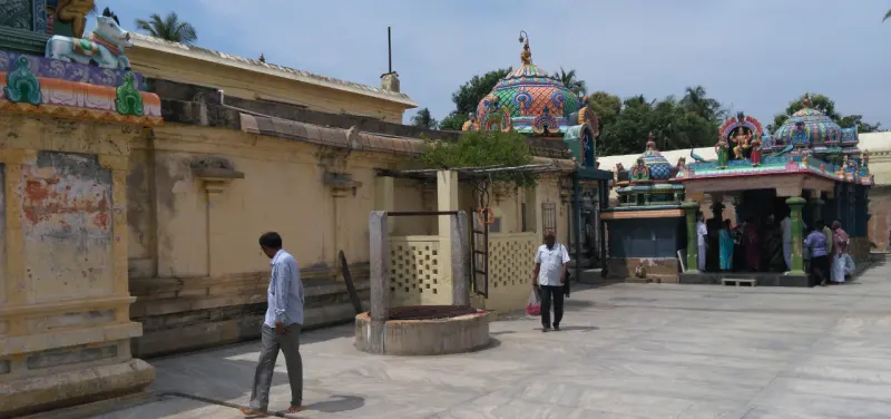 A low stone well in a paved courtyard, two men walking, shrines and domes behind