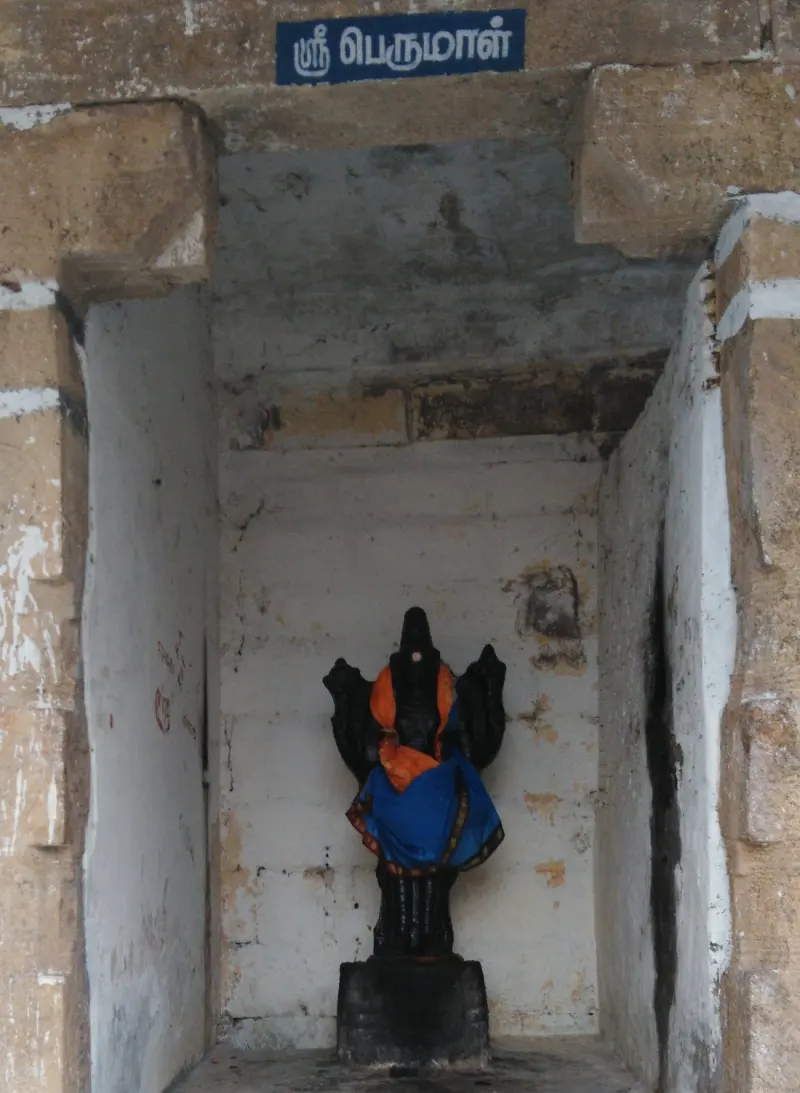 Narrow whitewashed niche holding a small dressed image on a black base, with a Tamil name board