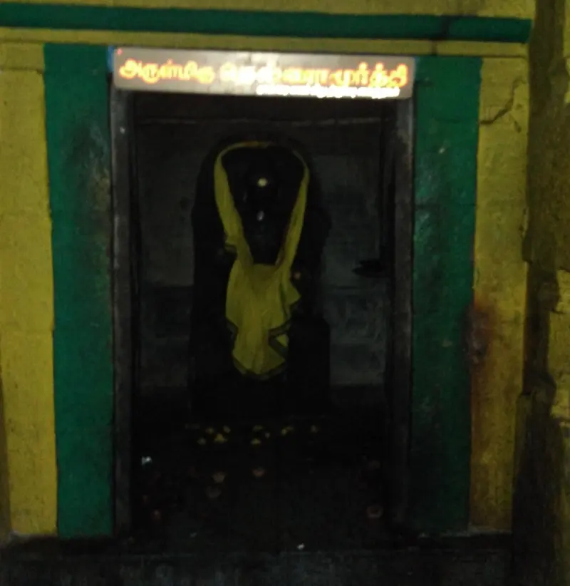 A dark doorway framed in green and yellow shows a figure draped in yellow cloth, under a lit Tamil sign.