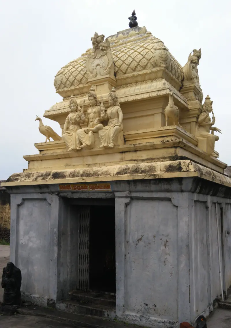 A grey shrine with a domed roof, three seated carved figures and carved peacocks, with a name board above the door.