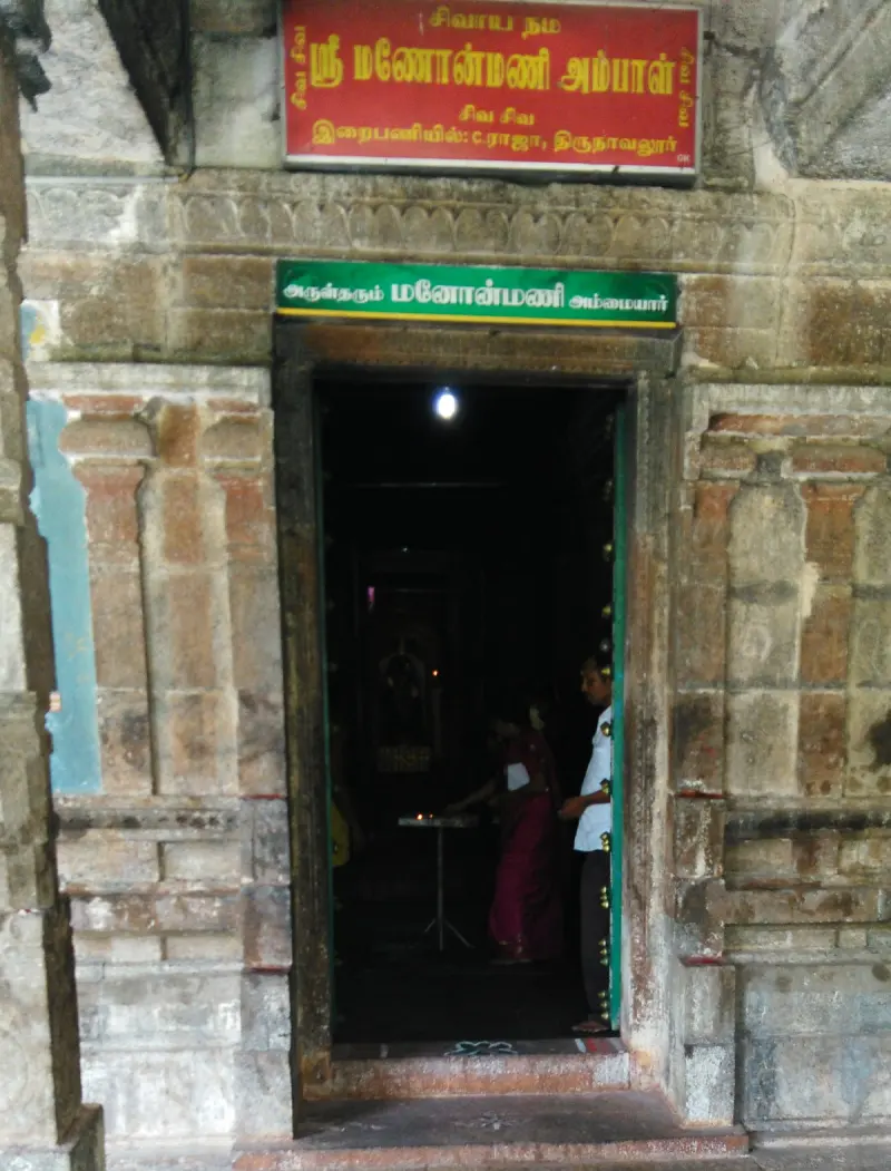 A dark doorway with Tamil name boards above it; a devotee and a man in white stand inside by a lit lamp.