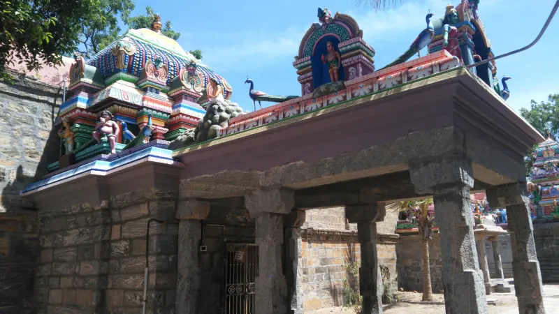 A stone mandapam with painted peacocks and figures on the roof, and a domed shrine above at right.