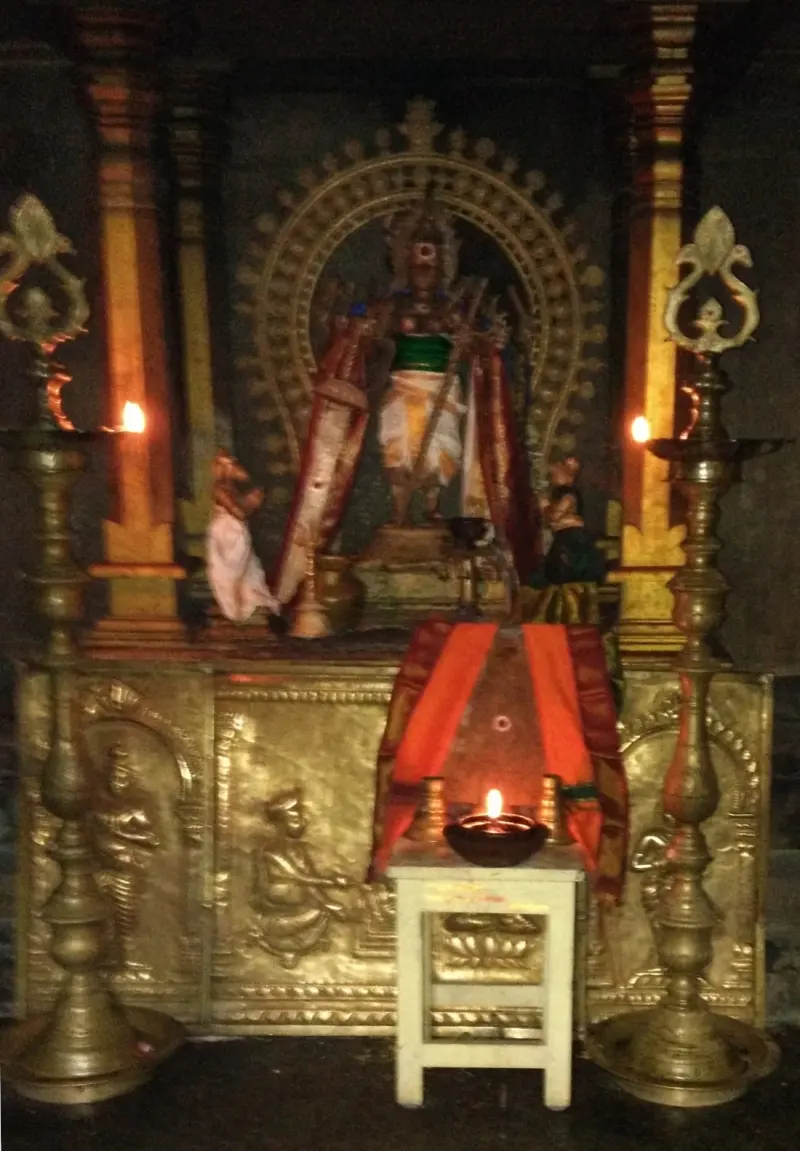 A standing idol in a robe before an arched frame, between two tall brass lamps, with a lit lamp in front.