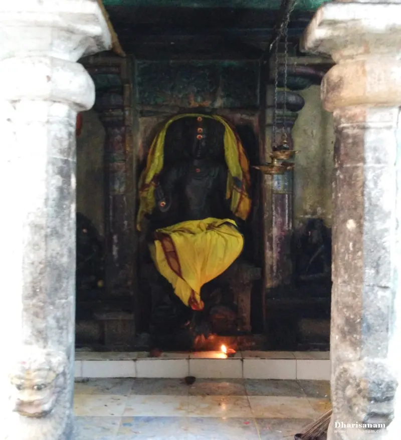 Dark stone seated deity draped in yellow cloth in a shrine between two pillars, with a lit lamp below