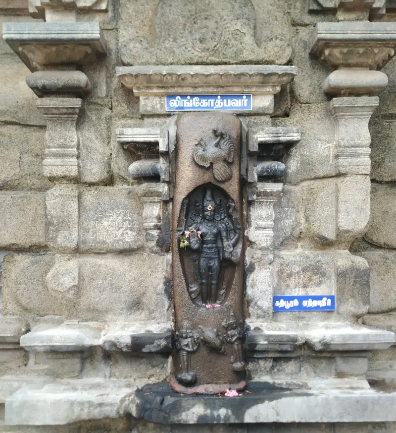 A dark carved figure in a tall niche on a stone wall, with a blue name board above reading Lingothbavar in Tamil.