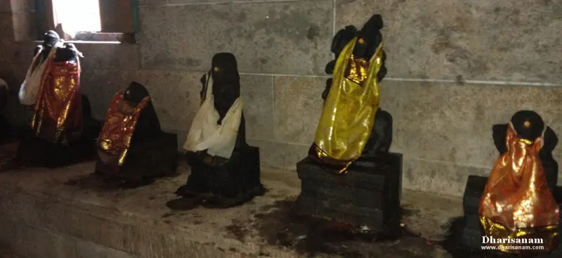 A row of black stone idols dressed in red, gold and white cloths on a low platform against a stone wall
