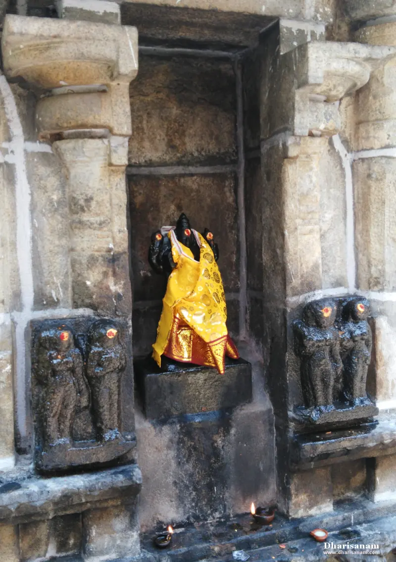 A dark stone Vinayakar draped in yellow cloth in a stone niche, with carved figures on both sides and oil lamps below