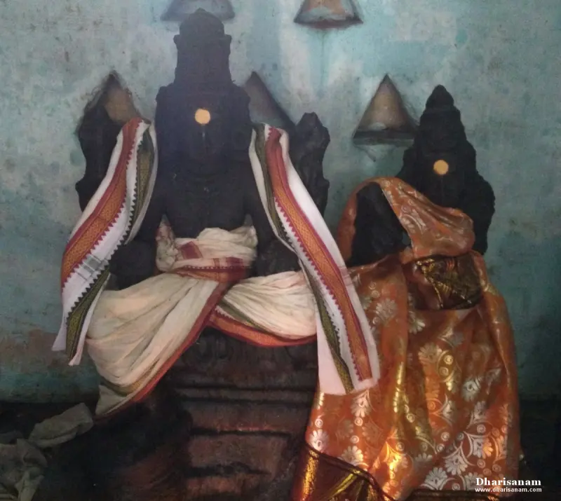 Two dark stone idols, one draped in white and the other in orange silk, against a pale blue wall