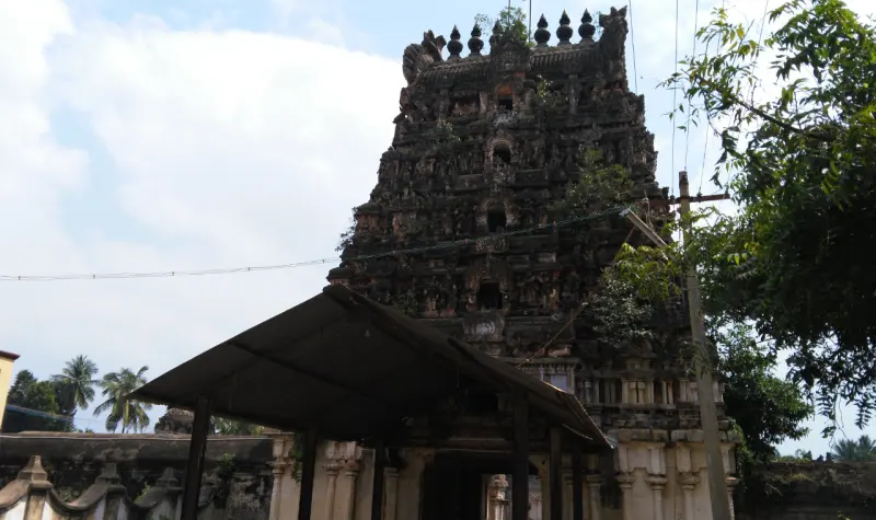 A dark weathered tiered tower with sculpted levels and plants on it, seen from under a roofed shelter.