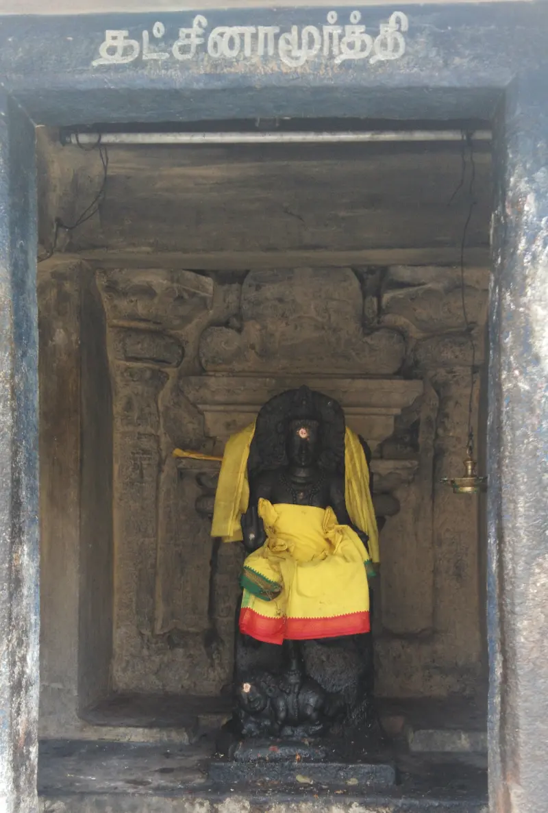 A dark seated idol draped in yellow under a Tamil name board