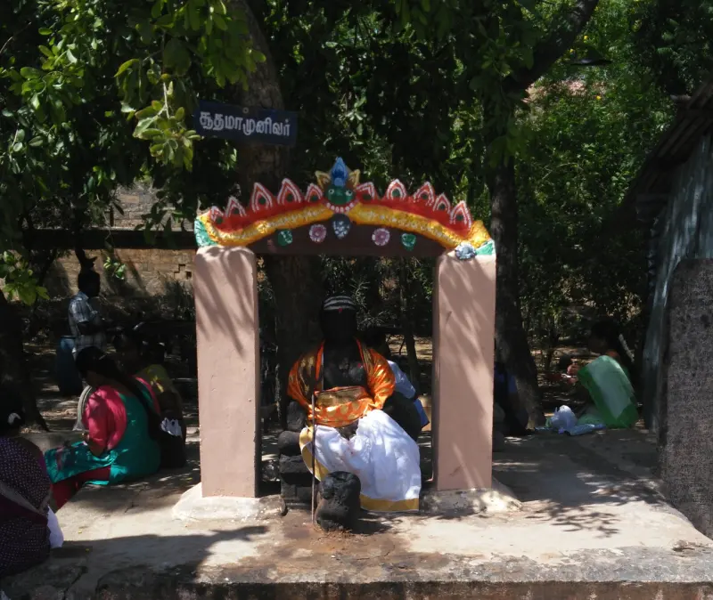A seated figure in orange and white cloth under a painted arch, with a Tamil name board above it.