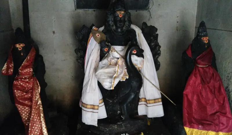 A dark stone figure in white cloth with a spear stands before a peacock, flanked by two women in red.