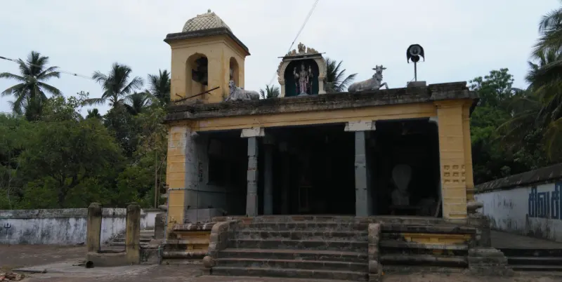 A yellow pillared hall with steps, a deity figure and a bull on its roof, and coconut palms behind.
