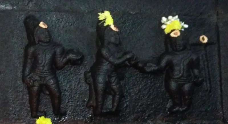 A black stone relief of three standing figures, two of them reaching toward each other, with yellow and white flowers