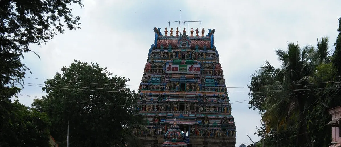 A tall, colourfully painted gopuram with rows of sculptures, seen between trees under a cloudy sky.