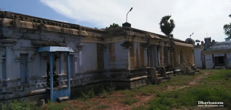 Long weathered stone corridor beside a grassy yard with a small blue shrine in front and palm trees behind
