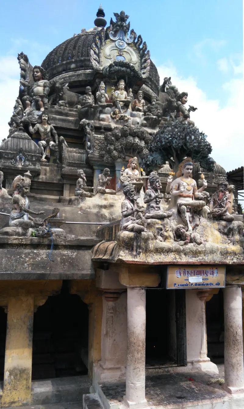A domed shrine roof carrying many stone and painted figures and a leafy tree sculpture, above a porch with a sign.