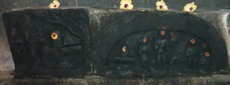 Weathered stone panels carved with small figures and a lingam, with hibiscus flowers along the top.