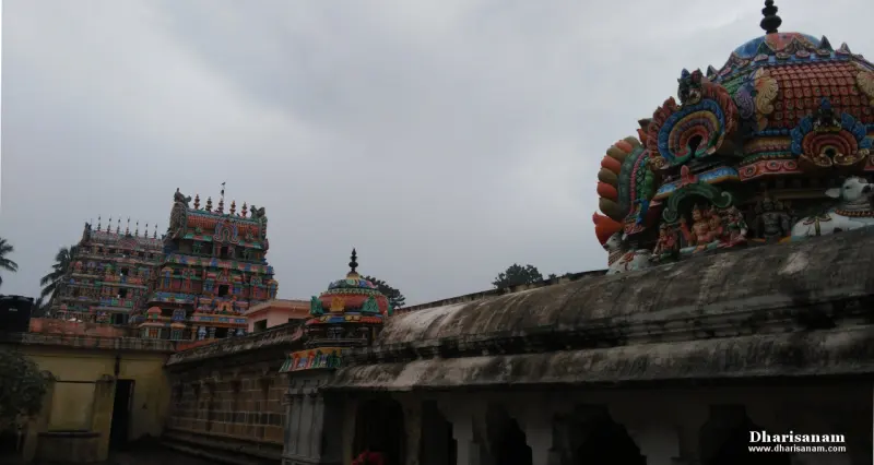 Colourful shrine roofs above a weathered wall, with two gopurams in the distance under a grey sky