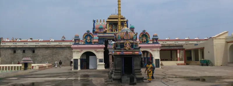 A golden flag post and a painted shrine in a wet courtyard, with a woman walking past and a tower behind