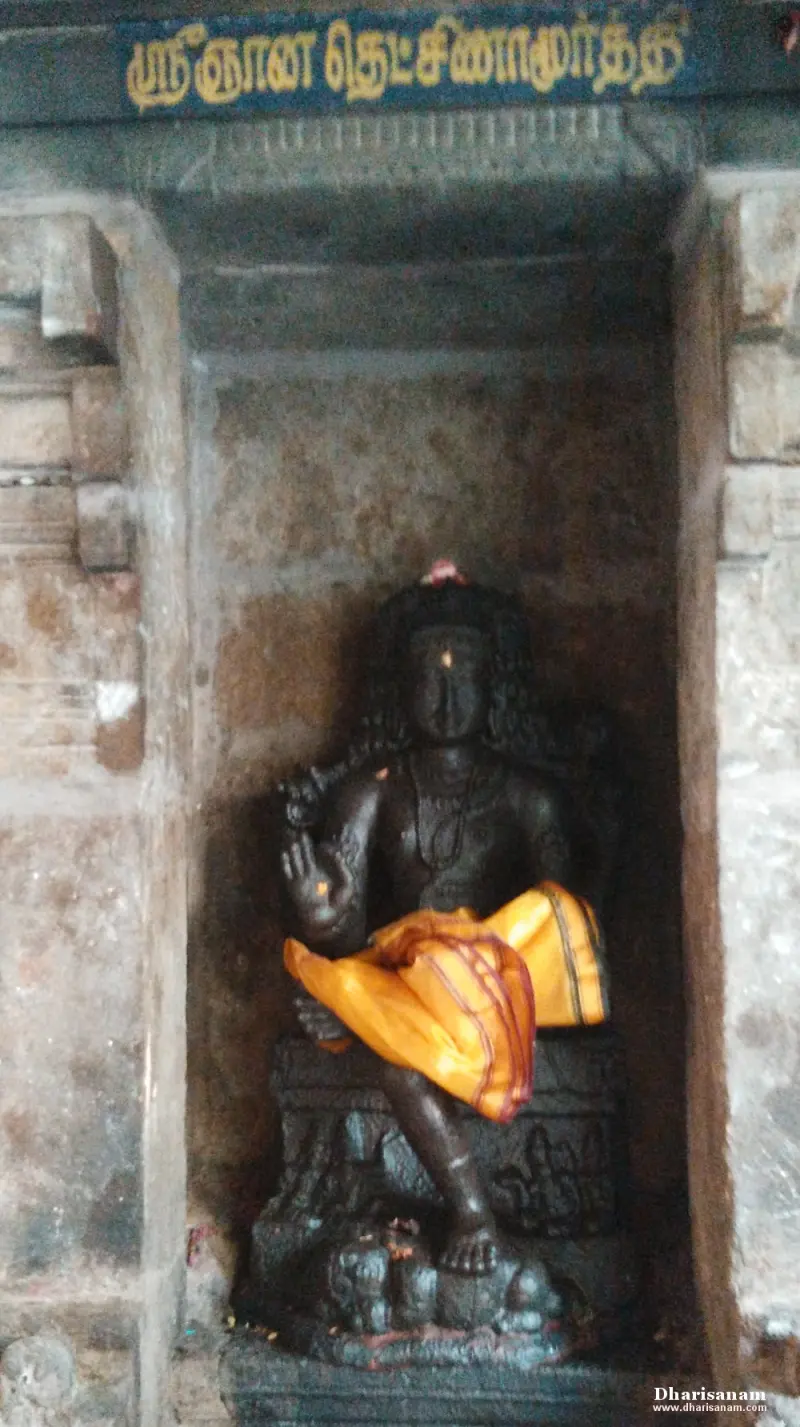 A dark stone idol seated in a niche with one hand raised and a yellow cloth over the lap, a Tamil name board above