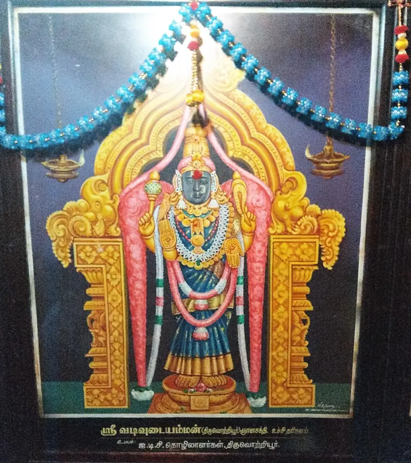A framed picture of a standing goddess with golden arch and pillars, with a garland and two lamps