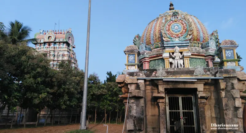 A shrine with a colourful domed roof and a gateway tower behind trees, under a blue sky