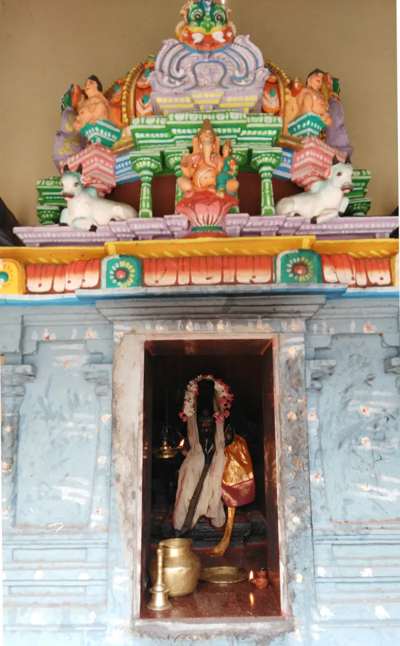 A pale blue shrine front with a painted Vinayakar and white bulls above, and a lit figure inside