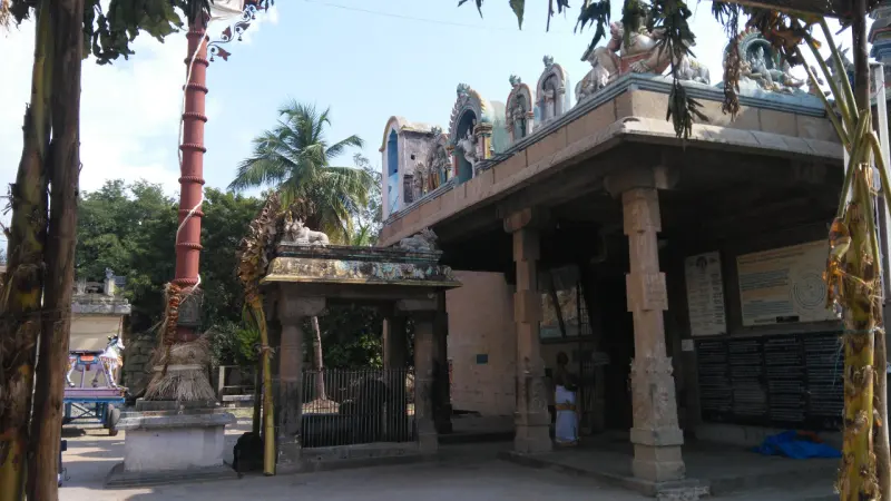 A tall red flagstaff tied with leaves beside a small stone mandapam, with a shrine roof and palm trees behind