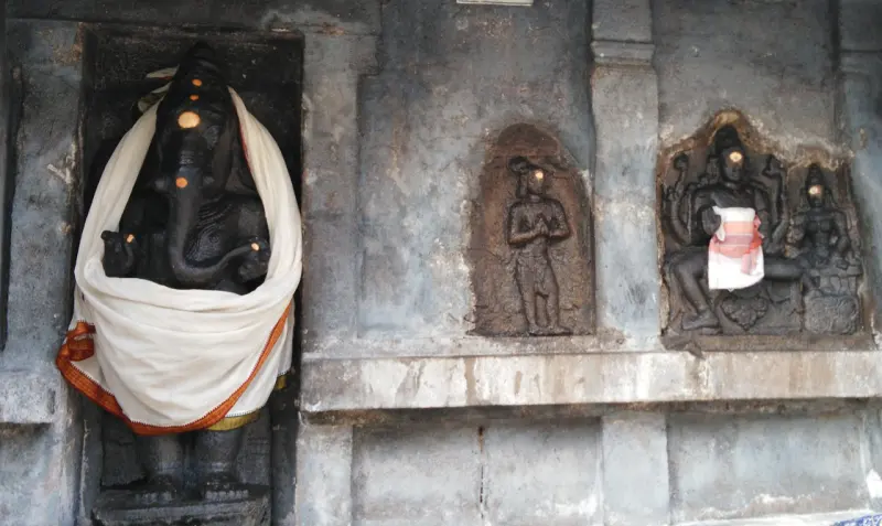 A black stone Ganesha draped in a white cloth beside two stone reliefs set into a grey wall