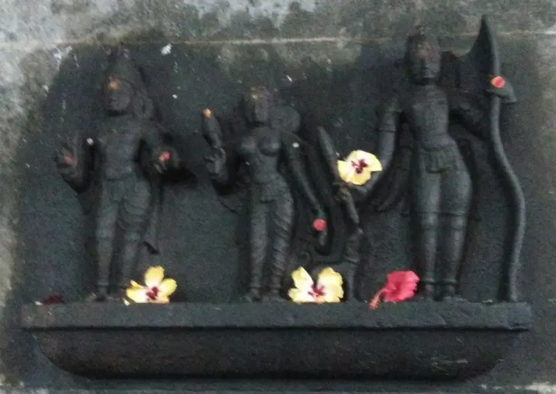 Dark stone relief of three standing figures, the one on the right holding a bow, with flowers at their feet.