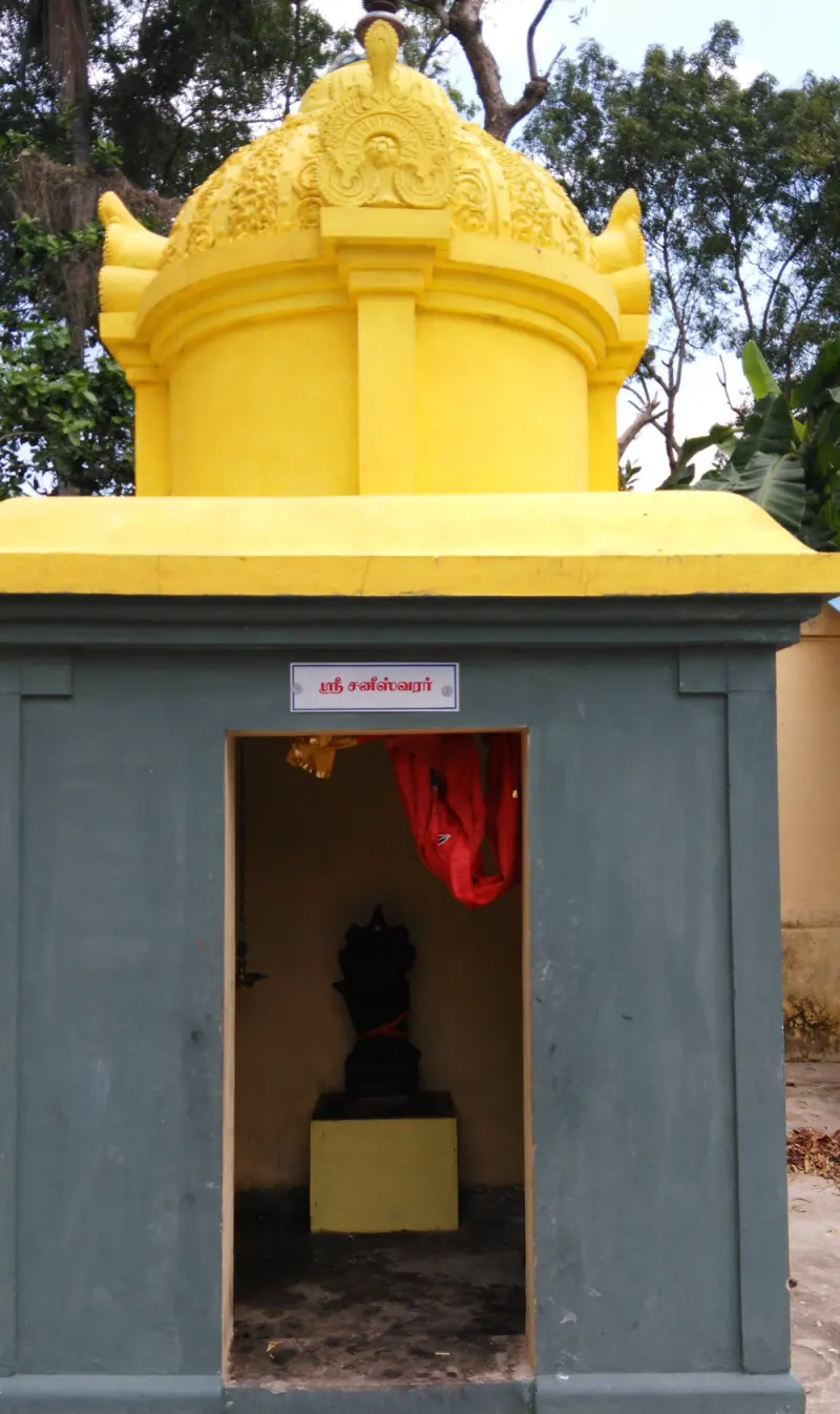 A small grey shrine with a yellow dome and a name plate; a dark idol with a red cloth stands on a yellow base