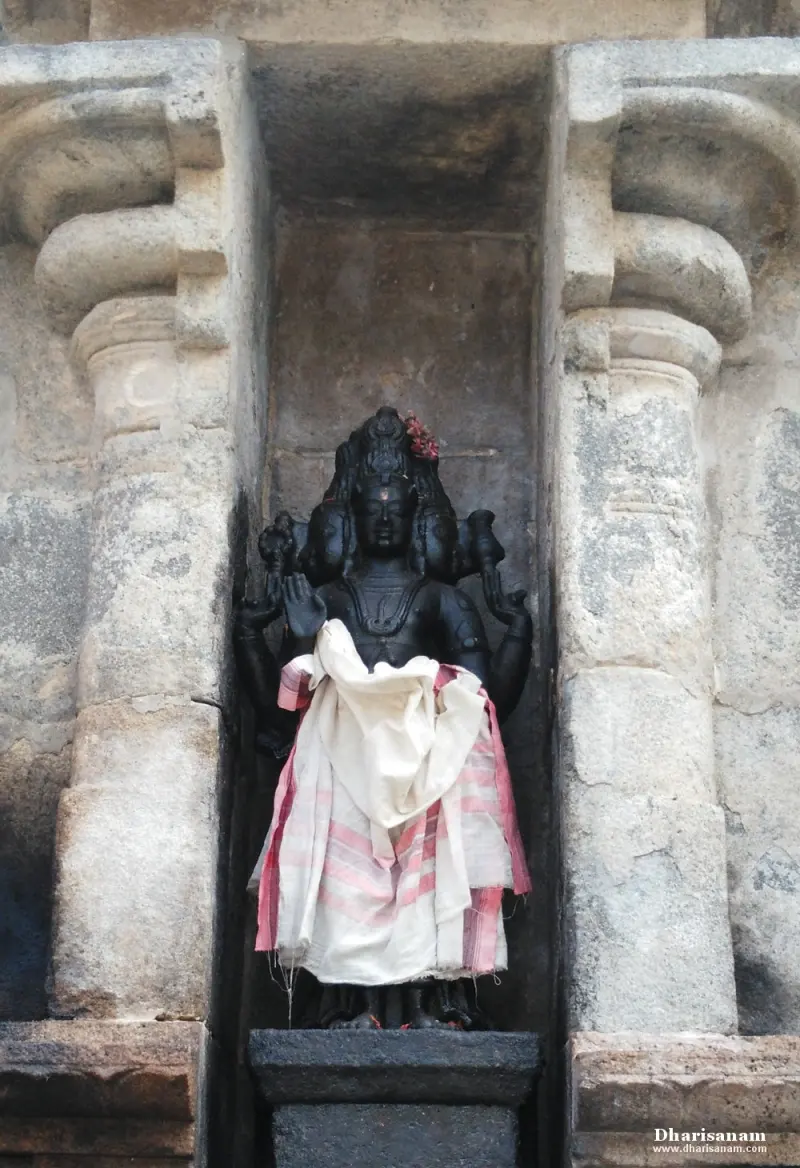 A dark idol with several heads and raised hands, wrapped in white cloth, stands in a stone niche