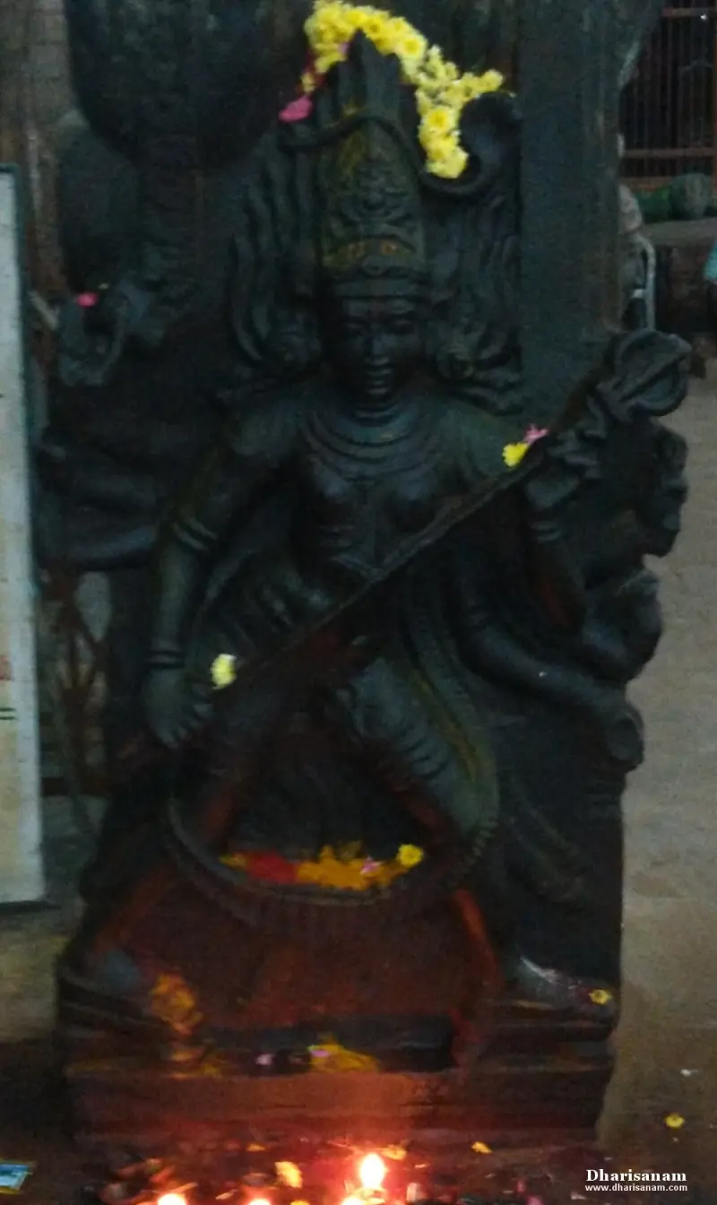 A dark stone idol of a goddess with many arms, a flower garland on her head and lamps lit before her