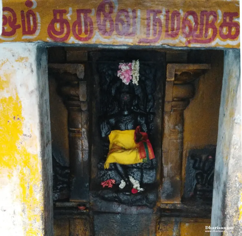 A dark stone idol wearing a yellow cloth, seated in a niche with flowers placed before it