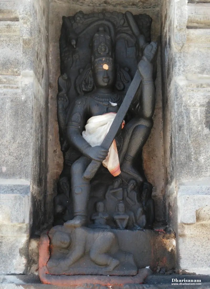 A dark stone goddess holding a long weapon, standing on a figure at her feet, in a wall niche.