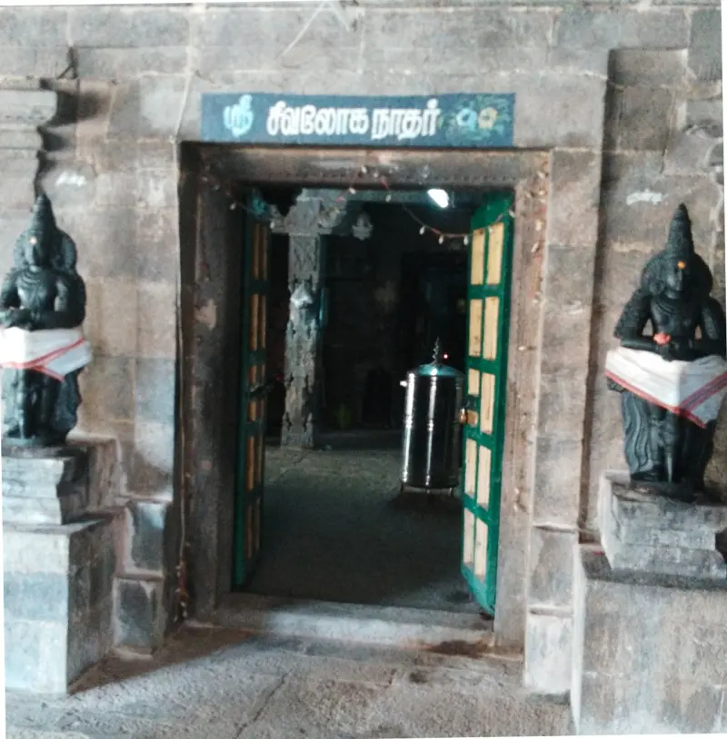 A stone doorway with a green door and a Tamil name board above it, flanked by two dark guardian figures in white cloth.