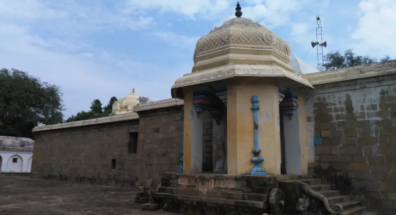A small domed mandapam on pillars stands at the corner of the stone wall, with steps