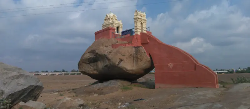 A large boulder in a dry riverbed with a red-painted wall, staircase and two small towers on top
