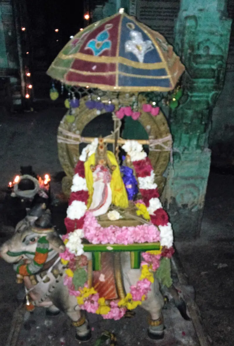 A small deity image covered in flower garlands sits on a stand under a coloured canopy, with oil lamps nearby.
