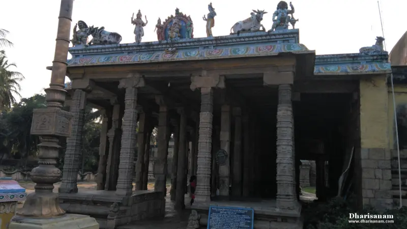 An open mandapam with carved pillars and painted figures along the roof, a flagstaff at left
