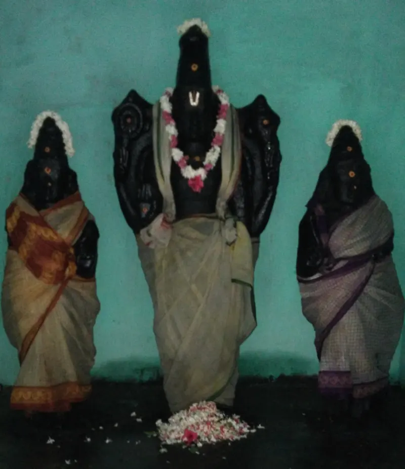 A tall dark figure with a garland, flanked by two figures in saris, in front of a green wall.