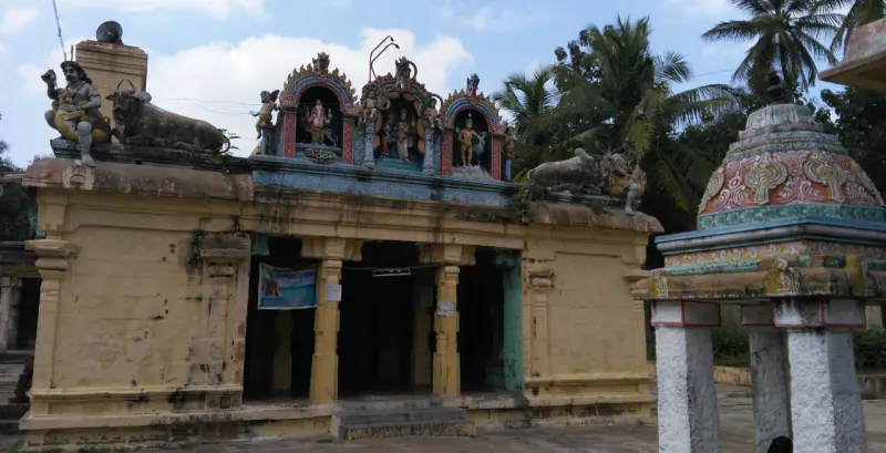 A yellow mandapam with painted figures and bulls on its roof, with a small domed shrine in the right foreground.