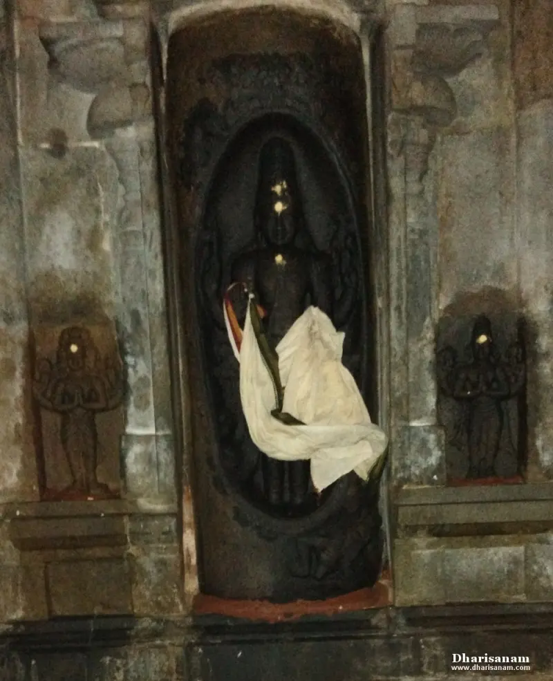 A dark stone standing figure in an oval niche with a white cloth, and a small praying figure on each side