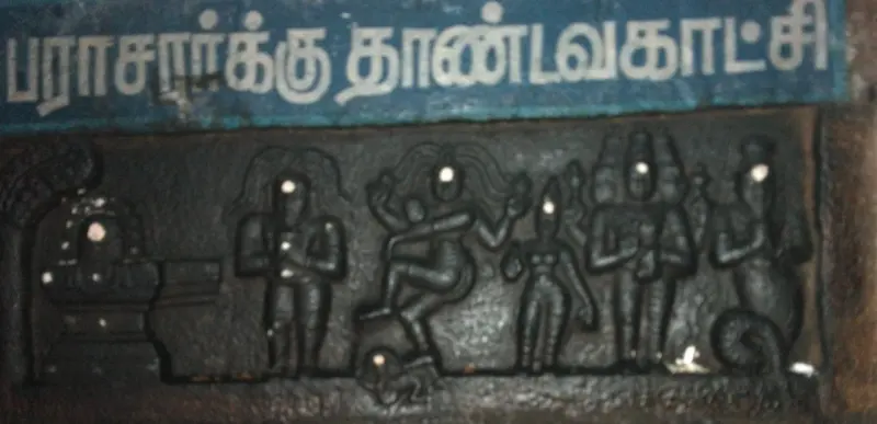 A dark stone relief with a dancing figure, a Shiva lingam and other carved figures under a Tamil caption board.
