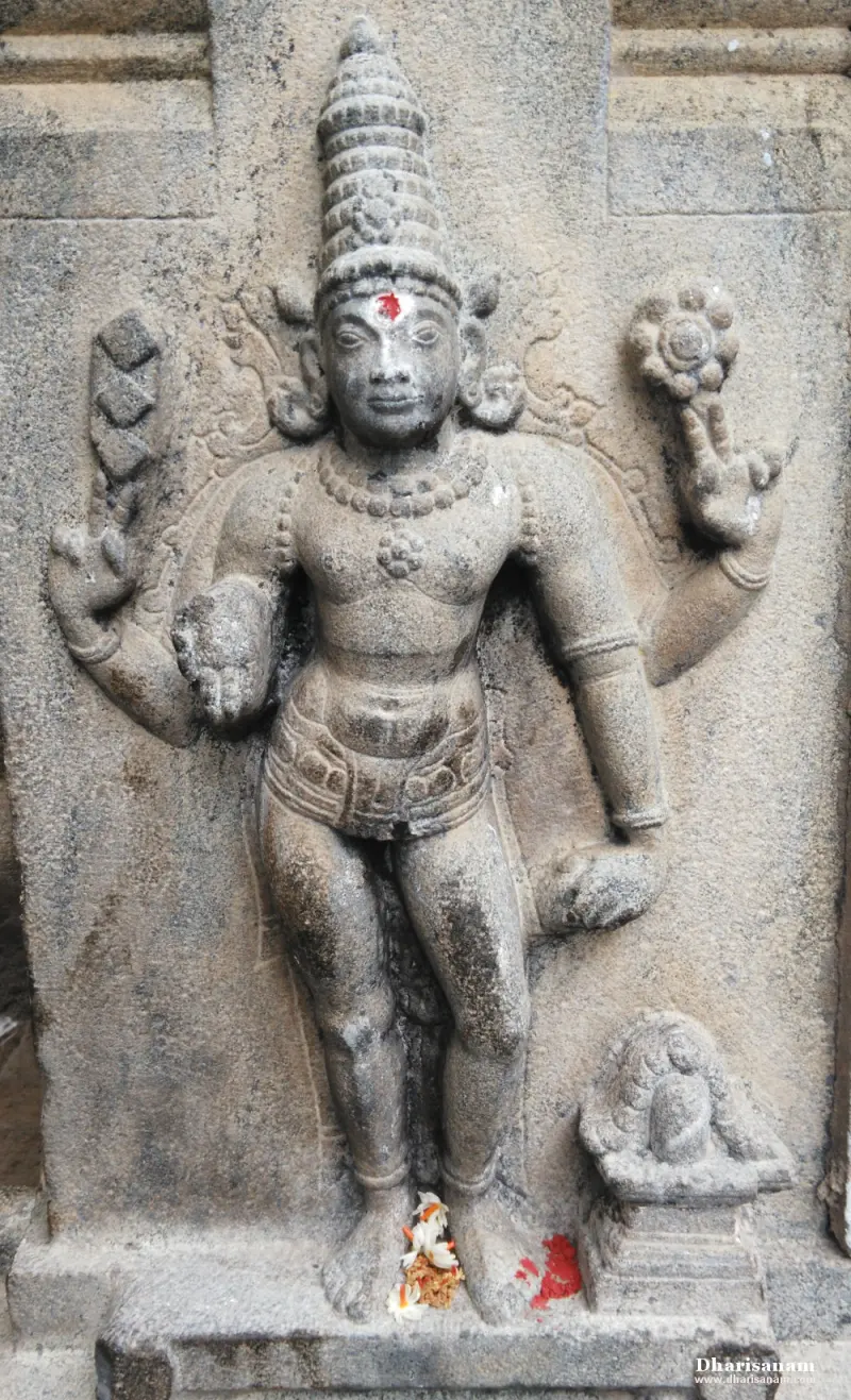 Standing four-armed figure with a tall crown, carved in stone, with flowers at its feet