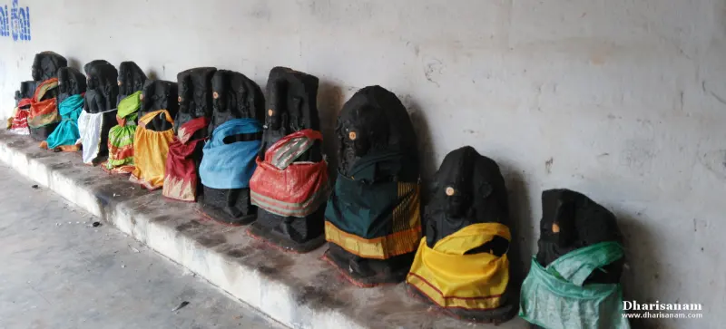 A row of small dark stone idols dressed in coloured cloths, seated along a stone ledge by a plain wall